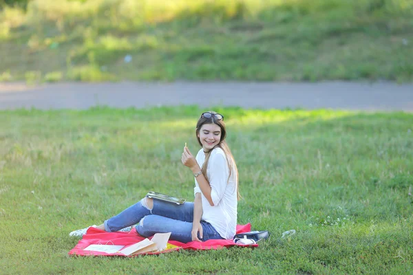 在一台笔记本电脑和吃快餐工作的年轻姑娘正坐在公园的草地上 — 图库照片