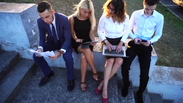 Бизнес-команда сидит рядом с центром офиса с документами, ноутбуком, планшетными технологиями против солнца — стоковое видео