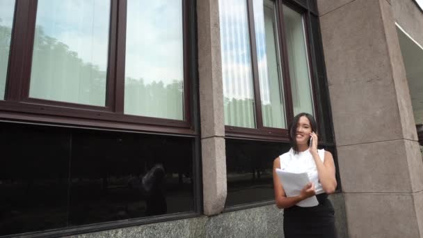 Fiatal gyönyörű üzleti nő női lány portré beszélni a telefont tartsa dokumentumok áll a háttérben irodaház szabadtéri — Stock videók