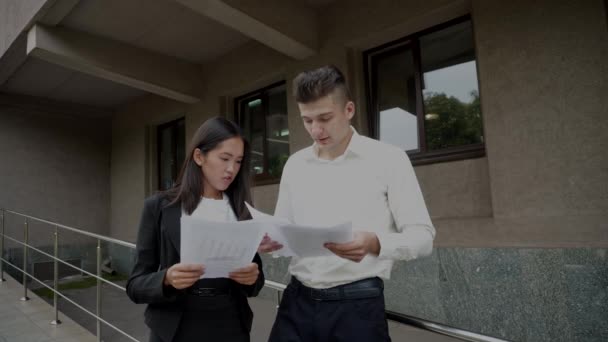 Joven hermosa asiática mujer de negocios hembra y caucásica macho mirar documentos charla en oficina edificio fondo — Vídeos de Stock