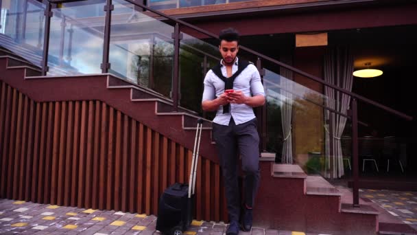 Arabska interesach mężczyzna wyjmuje telefon, wybiera numer i rozmowy na telefon komórkowy. Stojący w pobliżu schodów restauracji z czarna walizka. — Wideo stockowe