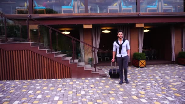 Jovem árabe empresário Guy caminha lentamente e rolar mala, sorrindo para a câmera no fundo do restaurante com a mão no bolso, e olhando ao redor . — Vídeo de Stock
