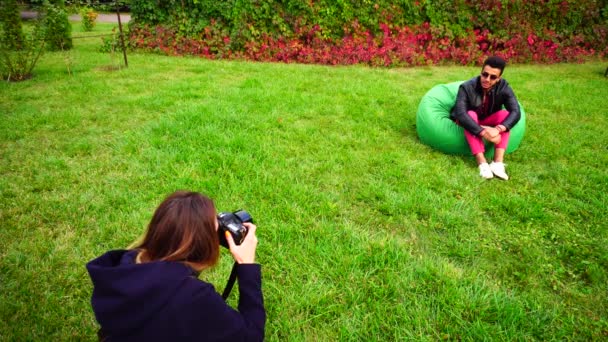 Hombre árabe encantador modelo posando y sonrisa en la cámara para el fotógrafo sentado en la silla en el jardín verde . — Vídeos de Stock