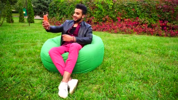 Lindo joven musulmán sosteniendo el teléfono en las manos, sonriendo, decidió llamar y sentarse en la silla en el jardín al aire libre . — Vídeos de Stock