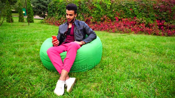 Piękny młody arabski człowiek trzyma w ręce telefon komórkowy, wzywa się Skype i siedzi w fotelu w ogrodzie na zewnątrz. — Wideo stockowe