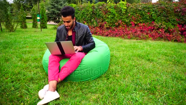 Empresario musulmán joven exitoso se sienta con el ordenador portátil en la silla, escribiendo en el teclado, mira a la pantalla y trabajando en línea, descansa en el jardín al aire libre . — Vídeo de stock