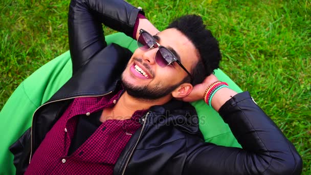 Retrato de jovem árabe homem masculino, estudante mente na cadeira, sorrindo, olhando para o céu, meditar no fundo da grama verde no jardim ao ar livre . — Vídeo de Stock