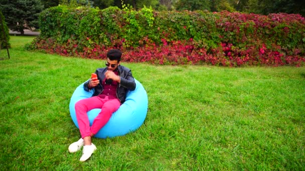 Muslim Guy, Empresario utiliza Smartphone y sostiene el teléfono en la mano, corrige el cabello y se sienta en la silla en el fondo del césped en el jardín al aire libre . — Vídeos de Stock