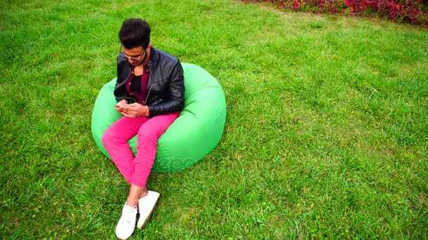 Empresario masculino musulmán majestuoso saca teléfono y responde llamada, hablando en el móvil, sonriendo y sentado en la silla en el jardín al aire libre . — Vídeos de Stock