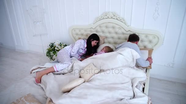Ребенок не слушает, но пара пытается спать, лежа на кровати в белом интерьере — стоковое видео