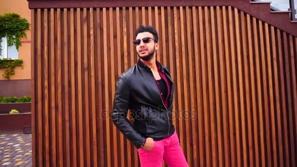 Bonito jovem árabe cara posando para câmera e endireita jaqueta ou cabelo, sorrisos e olha ao redor no fundo das escadas ao ar livre . — Vídeo de Stock