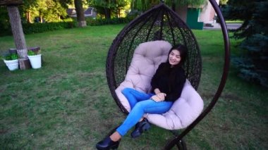 Güzel kadın bir ayağı, görünüyor ve gülümsüyor, kameraya poz koyarak askıya sandalyede rahatça oturur. Park açık havada sallanan sandalye.