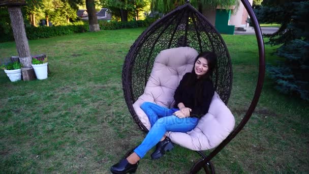 Pretty Woman Sits Comfortably in Chair Suspended by Putting One Foot, Looks and Smiles, Posing at Camera (dalam bahasa Inggris). Goyang Kursi di Taman Outdoors . — Stok Video