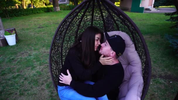Garçon et fille assis ensemble dans une chaise suspendue et s'embrassant, souriant et parlant dans un grand parc en plein air . — Video