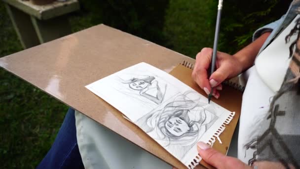 La hembra sostiene el lápiz gris en la mano, hace bocetos y esboza en un pedazo de papel y se sienta en el parque al aire libre . — Vídeo de stock