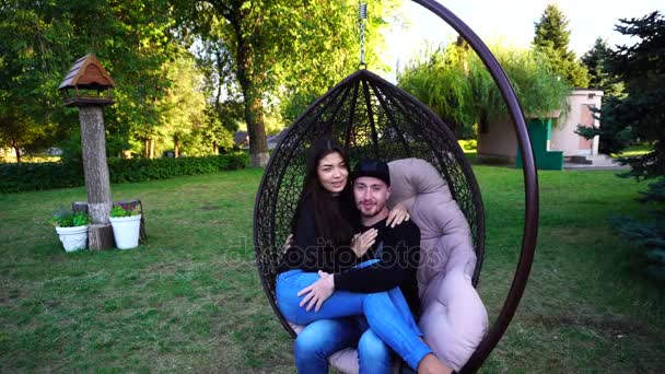 Menino e menina sentados juntos em cadeira suspensa, posando na câmera e sorrindo em grande parque ao ar livre . — Vídeo de Stock