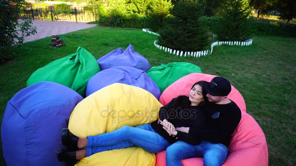 Jonge vriend en vriendin zitten en praten in de veelkleurige fauteuils en glimlachend, knuffelen andere buiten in Park. — Stockvideo