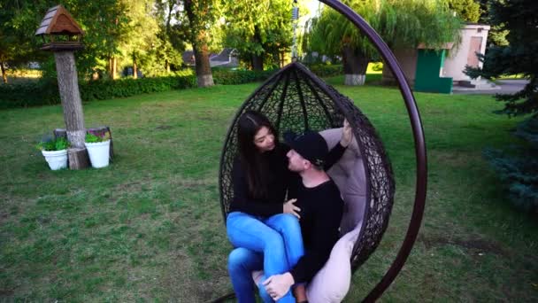 Recién casados sentados juntos en silla de mimbre y abrazándose mutuamente y hablando en un gran parque al aire libre . — Vídeos de Stock