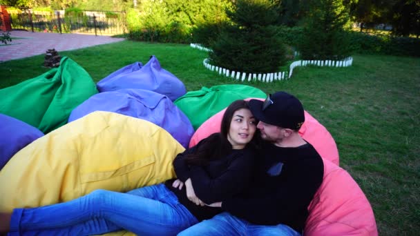 Casal apaixonado abraçando uns aos outros e conversar em poltronas multicoloridas, sorrindo e sentado ao ar livre no parque . — Vídeo de Stock