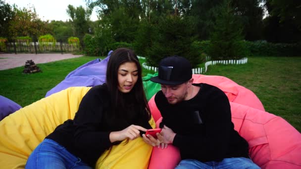 Atraente jovem casal amoroso decidiu ter trabalho ou descanso e usando Smartphone no parque, sentado em cadeiras confortáveis ao ar livre . — Vídeo de Stock