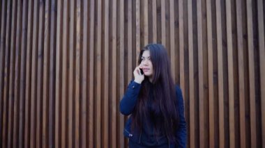 Güzel esmer genç yetişkin kız el telefon ve sohbet eden, tutan gülümseyerek ve konuşurken gülüyor, günde duvarının yakınında duran zaman açık havada.