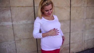 Sevimli genç hamile kadın yavaşça dokunmadan Belly ve içine kamera, duvar arka planı açık havada gündüz ayakta poz.