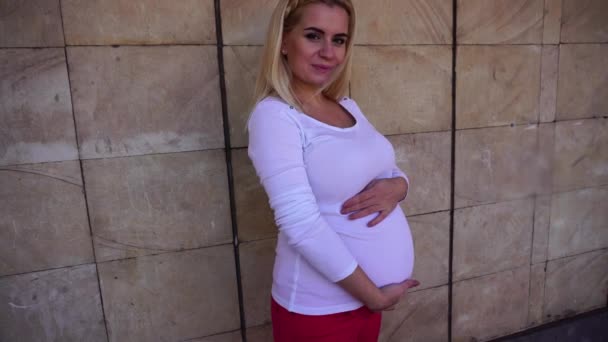 Jolie femme enceinte blonde caressant doucement le ventre et souriant, posant à la caméra et debout contre le fond du mur en plein air en journée . — Video