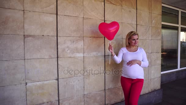 Charmante blonde va devenir mère, regardant dans la caméra sourit et tient à côté de ballon, Poses pour photo sur fond de mur de pierre à l'extérieur . — Video