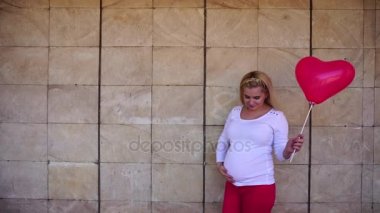 Sevimli genç yetişkin Bayan balon el, bakar, gülümser ve kamera tutan bebek jest ve duvar açık arka plan üzerinde duruyor gösterir bekliyor.