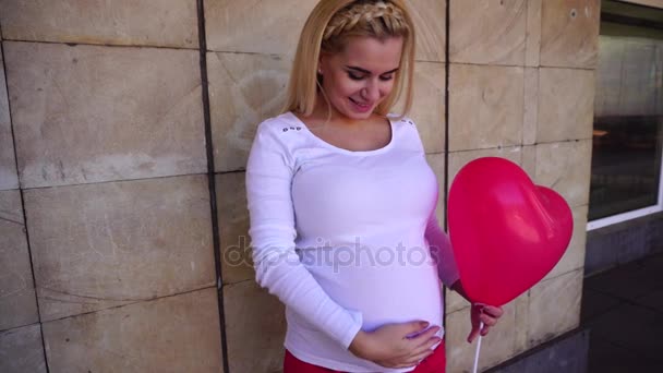 Chica embarazada rubia acariciando suavemente el vientre, sonriendo y posando en la cámara y sosteniendo en globo de mano contra el fondo de la pared al aire libre durante el día . — Vídeos de Stock
