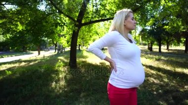Çarpıcı genç hamile kadın yavaşça dokunmadan göbek ve yeşil güneşli Park açık havada gündüz ayakta rahatsızlık geri, hissediyor.