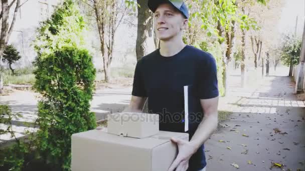 Postbote trägt Paket in Händen lächelnd in die Kamera auf dem Hintergrund von Bäumen entlang der Straße . — Stockvideo