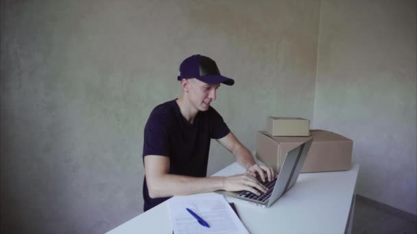 Deliveryman πληκτρολόγηση σε Leptop και πάρτε στυλό, γραπτώς σε χαρτί και κάθεται στο γραφείο κοντά σε κουτιά στο φόντο του τοίχου στο ταχυδρομείο. — Αρχείο Βίντεο