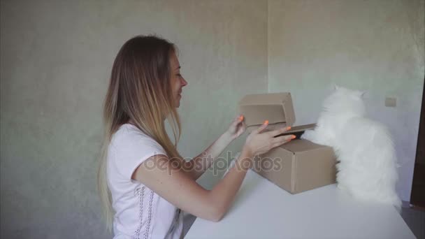 Chica se sienta a la mesa en la habitación, abre una pequeña caja cerca de su gato se sienta y mira a su alrededor . — Vídeos de Stock