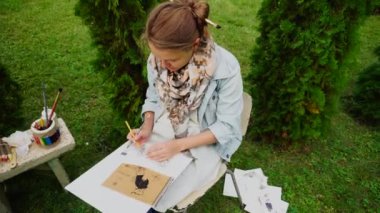 Kadın Illustrator eskiz kağıt üzerinde yere özetliyor ve kalemleri kullanarak ve elinde tutar. Kız parkta gündüzleri açık havada oturur.