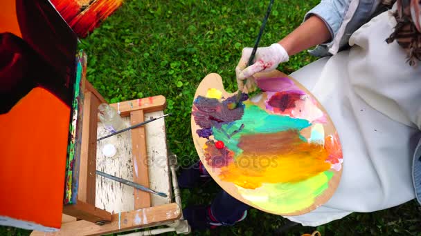 Mão pintor fêmea mistura cores para obter sombra desejada para pintar imagem e senta-se na cadeira atrás cavalete ao ar livre fora no parque . — Vídeo de Stock