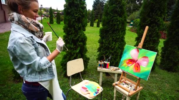 Linda mujer artista obras y soportes con pincel en la mano cerca del caballete, buscando y acabados pintura de pie en Green Park al aire libre . — Vídeos de Stock