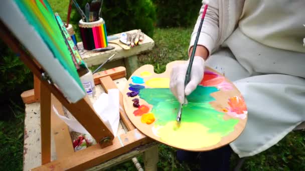 Hermosa artista mezcla amarillo con pintura verde y sentado en el parque cerca de bancos con suministros de arte al aire libre . — Vídeo de stock