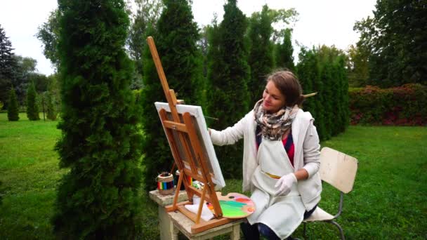 Impresionante artista mujer sentada en el caballete, dibuja en el parque en el fondo de árboles de coníferas al aire libre . — Vídeos de Stock