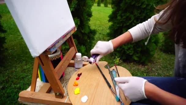 Művész lány gazdaság cső a festék, megnyílik, és tolja a festék a palettán, kint ül a festőállvány a Park. — Stock videók