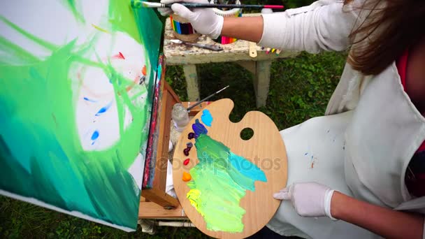 Lidová škola umění školačka barvy barvy v ruce drží Štětec pozadí a vést na plátně, paleta drží na nohou a sedí v parku venku.