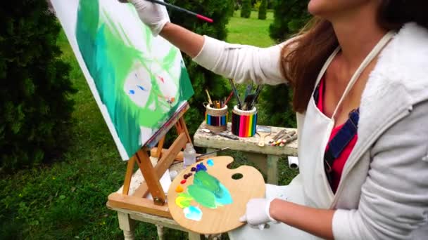 Atractiva mujer creativa sostiene cepillos y dibuja imagen en el parque sentado en el caballete al aire libre en el fresco día de otoño . — Vídeos de Stock