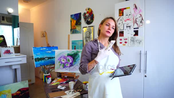 Дівчина художник тримає мобільний телефон і використання, розмовляє, пише ручку на дошці з листям і посміхається, стоїть в художній студії . — стокове відео