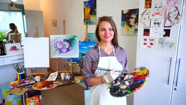 La colegiala de la escuela de arte mira a la cámara y en la paleta, sonríe y se encuentra entre el estudio para dibujar que paredes en imágenes . — Vídeo de stock