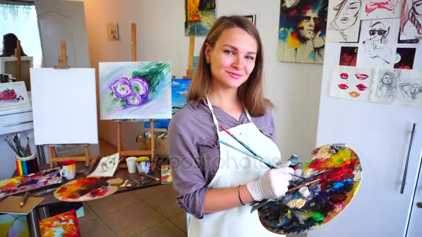 Jolie jeune professeur d'école d'art souriant à la caméra avec des brosses à la main, qui tient dans la palette dans le studio d'art accroché avec des croquis et des dessins . — Video