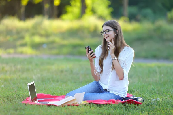 Jeune fille assise dans le parc, souriant et travaillant à son ordinateur portable, écoutant de la musique — Photo