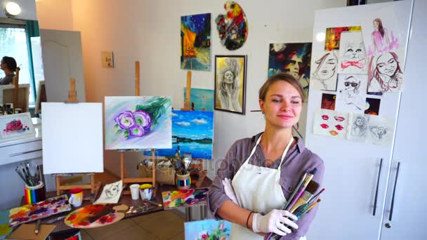 Hermosa artista se para frente a la cámara con los brazos cruzados delante de ella y sostiene en cepillo de mano en el estudio de arte . — Vídeo de stock