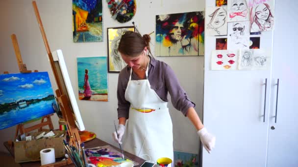 Bella ragazza che sceglie la pittura dell'artista su pennello e godendo della musica, sorridendo e ballando in Art Studio . — Video Stock
