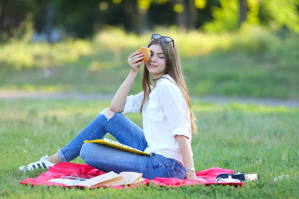 Giovane ragazza seduta sull'erba nel parco e lavora in un computer portatile e mangiare fast food — Foto Stock