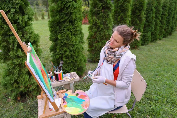 Chica artista pinta cuadro y se sienta en la silla a los lados del caballete de t —  Fotos de Stock